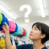 大人用の水泳帽はどこで買える？と悩む日本人女性が、お店で水泳帽を前に考えている様子。購入場所を探すイメージ。