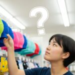 大人用の水泳帽はどこで買える？と悩む日本人女性が、お店で水泳帽を前に考えている様子。購入場所を探すイメージ。